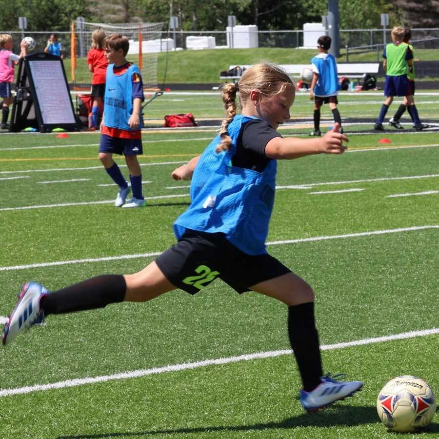 A camper in a blue bib winds up to strike the ball on the turf